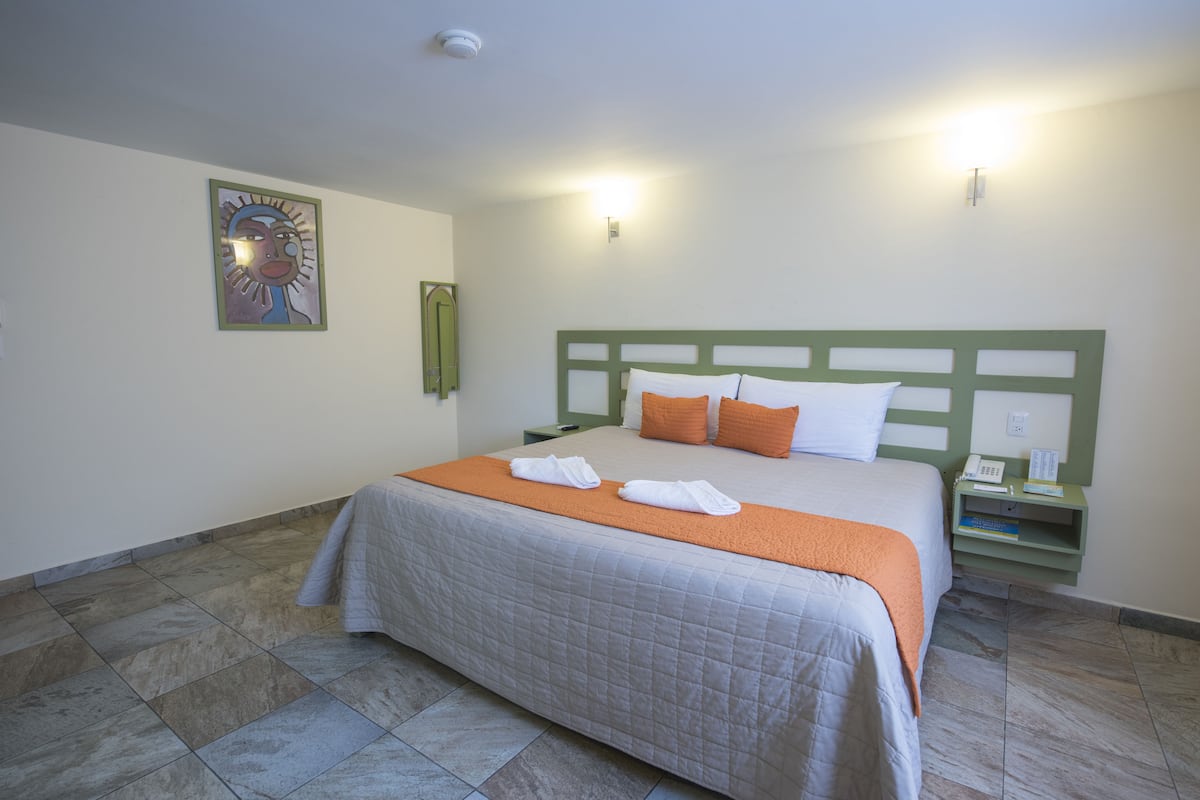 Una cama bien hecha con una manta y almohadas anaranjadas, un cabecero verde, una mesita de noche con una lámpara y una obra de arte colgada en la pared.