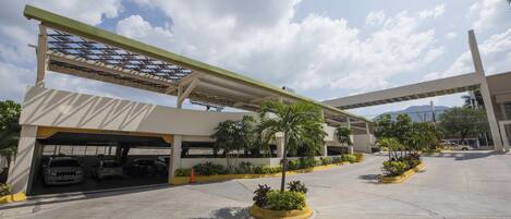 Un edificio moderno con estacionamiento techado, varios coches y jardineras ajardinadas.