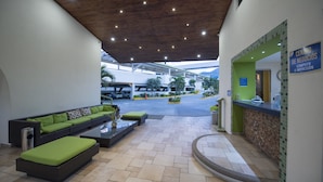 Un área de recepción con mostrador, zona de estar con sofá y reposapiés, y suelo de baldosas.