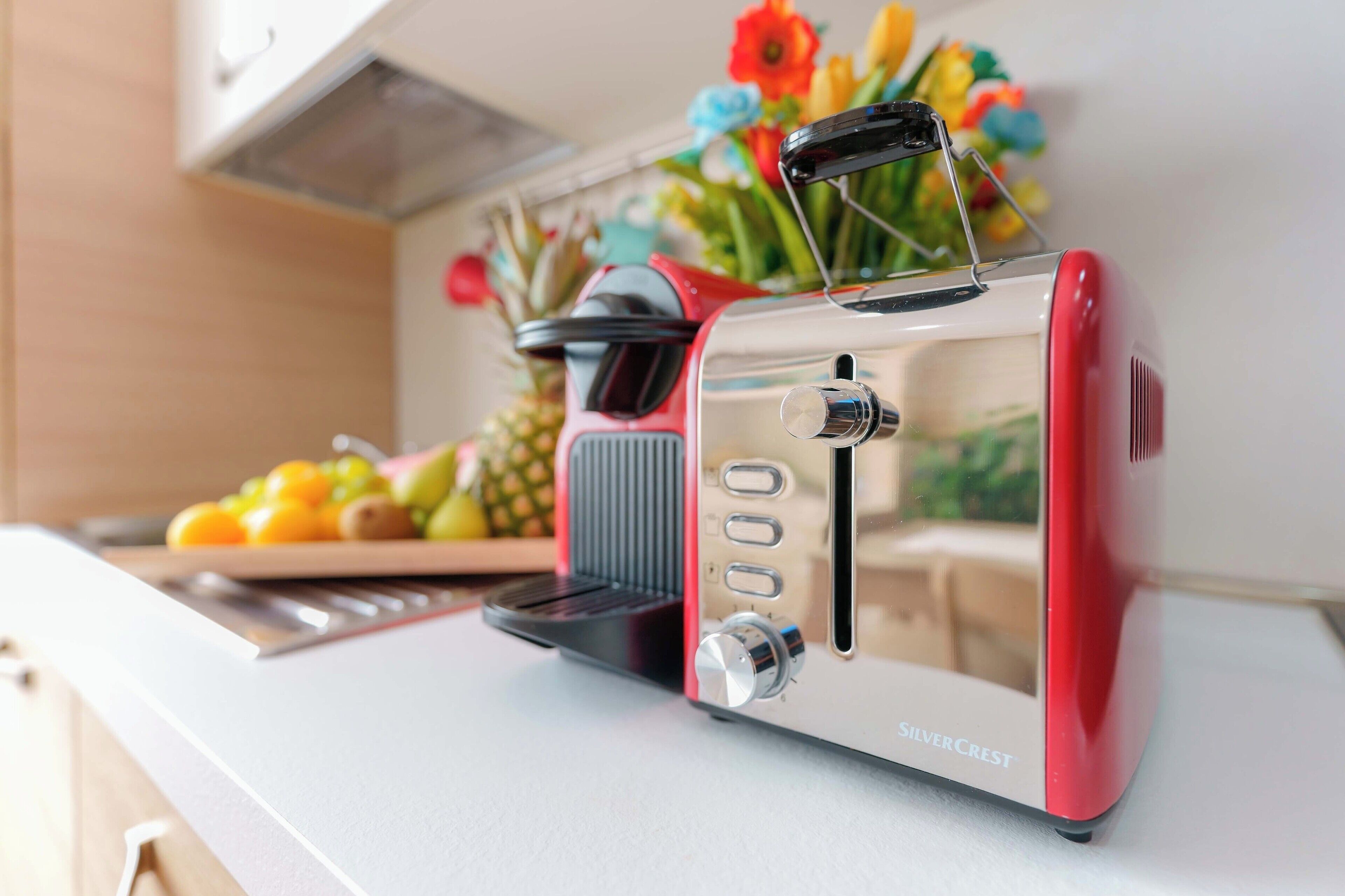 Une cuisine avec un grille-pain rouge et blanc posé sur le comptoir, un panier de fruits et un vase rempli de fleurs.