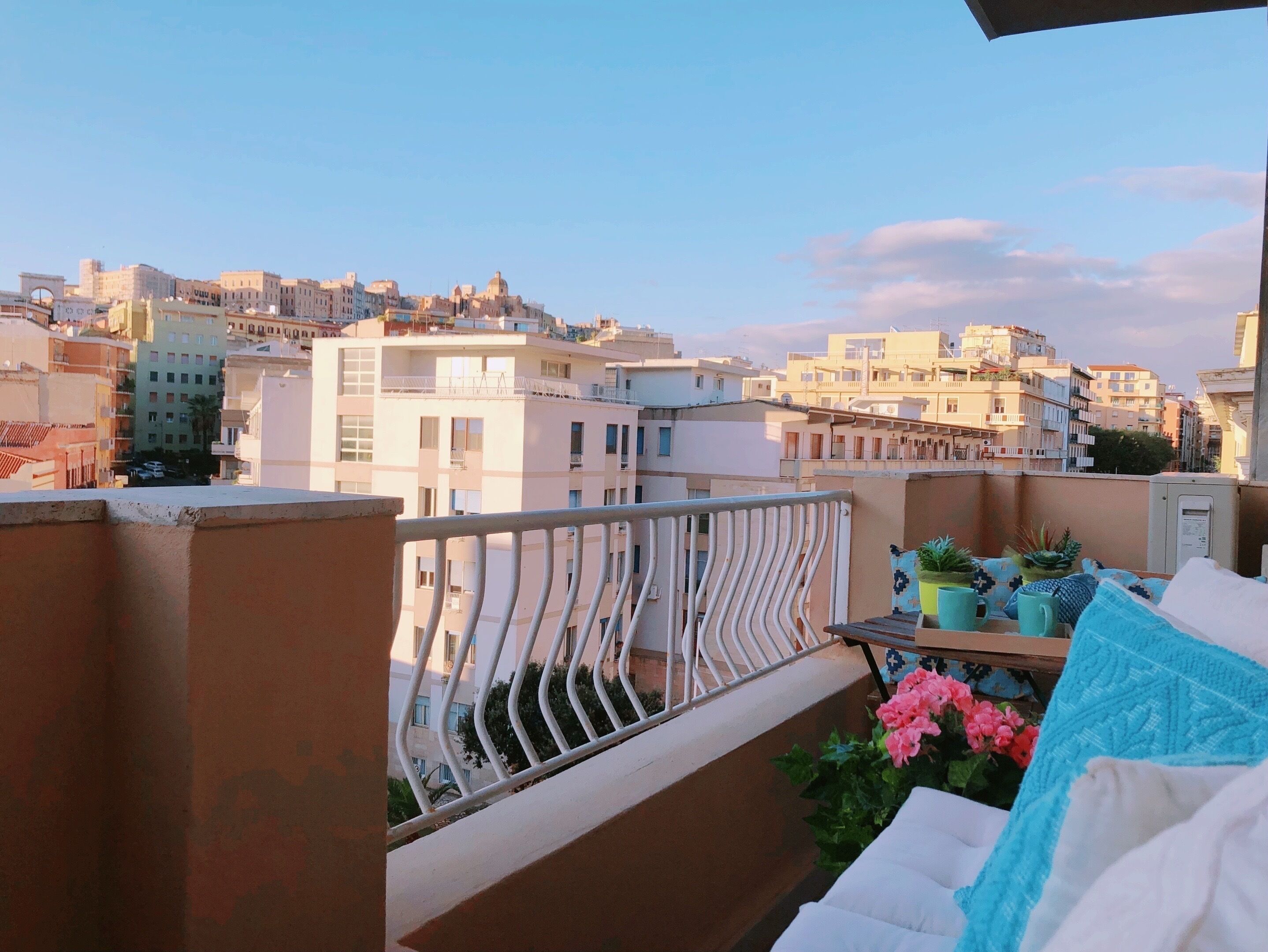 A balcony with a railing, potted plants, and a view of a cityscape with buildings.