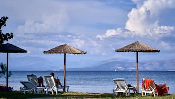 Eine Strandlandschaft mit Liegestühlen und Strandhütten aus Palmwedeln, mit Blick auf ein ruhiges Meer und ferne Berge.