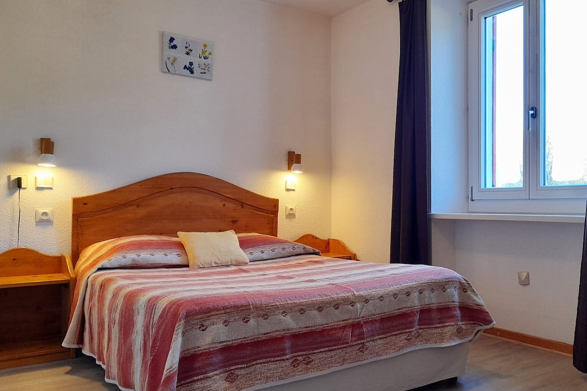 A bedroom with a wooden bed, two bedside tables, a nightstand, a window with curtains, and a wall-mounted light fixture.