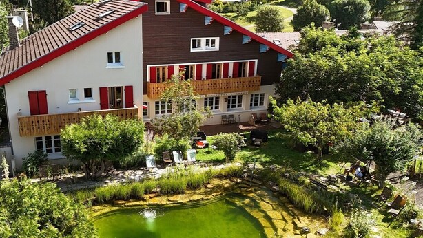 Un edificio de dos pisos con techo rojo, un pequeño estanque y follaje circundante.