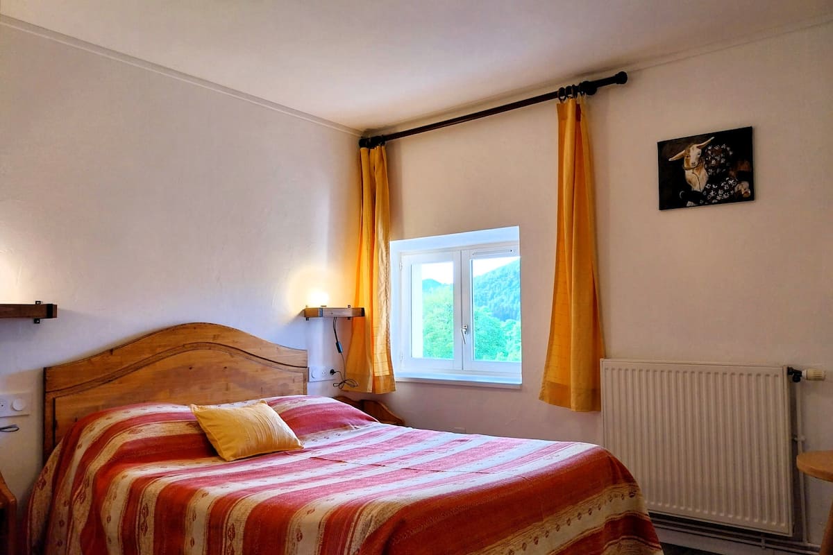 A bedroom with a wooden headboard, a bed with a striped bedspread, a window with curtains, a radiator, and a wall-mounted picture.