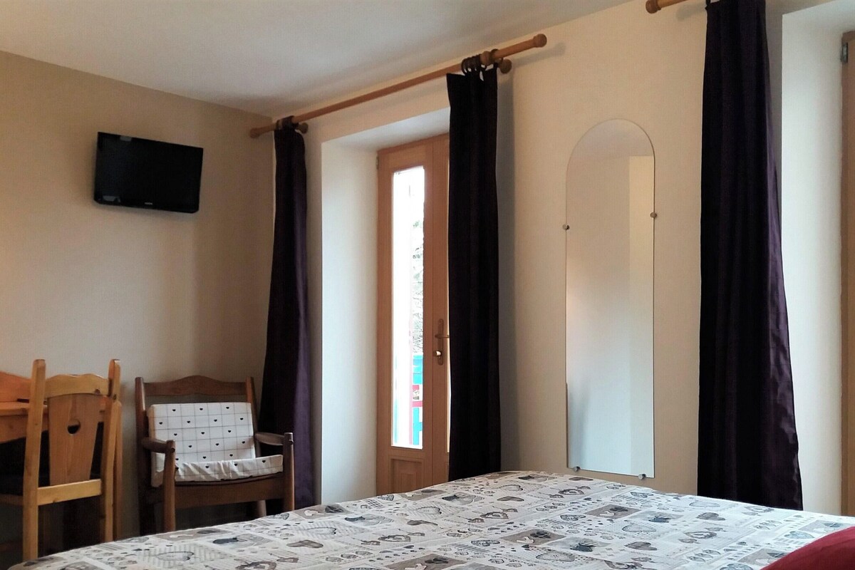 A bedroom with a bed, a chair, a desk, a television, and a door leading to a balcony.