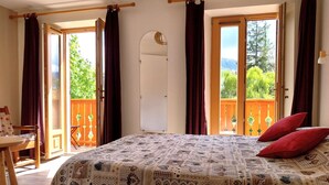 Un dormitorio con una colcha estampada, dos almohadas rojas, una silla de madera, una mesa y un balcón con vistas a la vegetación.