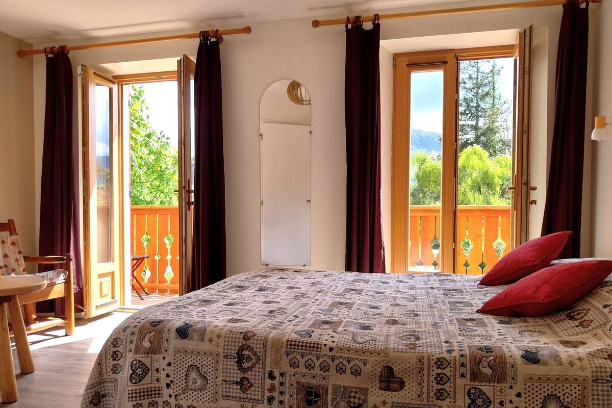 A bedroom with a patterned bedspread, two red pillows, a wooden chair, a table, and a balcony with a view of greenery.