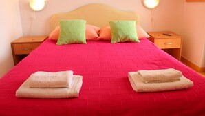 A neatly made bed with a red bedspread and two green pillows, two folded white towels, and a wooden nightstand with a remote control. - Guest House Tomanovic (Herceg Novi)