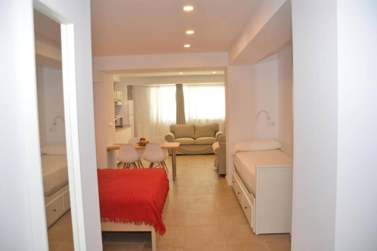 A compact living space with a bed, a red blanket, a dining area with chairs, a sofa, and a window with curtains.