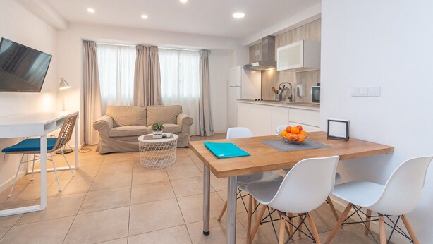 A modern living room with a sofa, a TV, and a dining area with a table and chairs. - Bet Apartments - Abastos (Valencia)