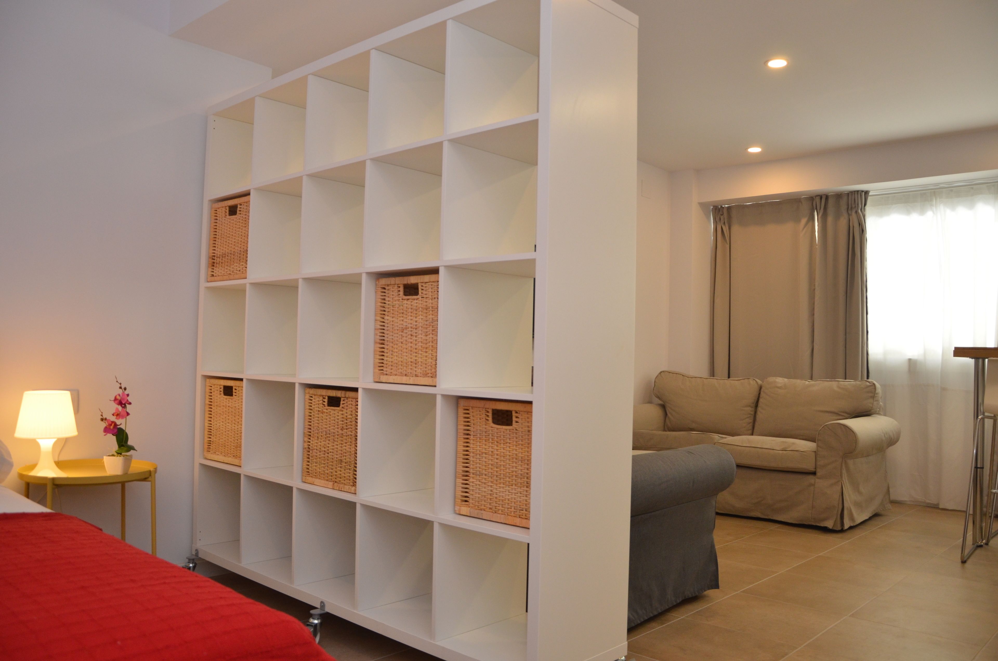 A hotel room with a bed, a sofa, a white shelving unit with baskets, a lamp, and a window with curtains.