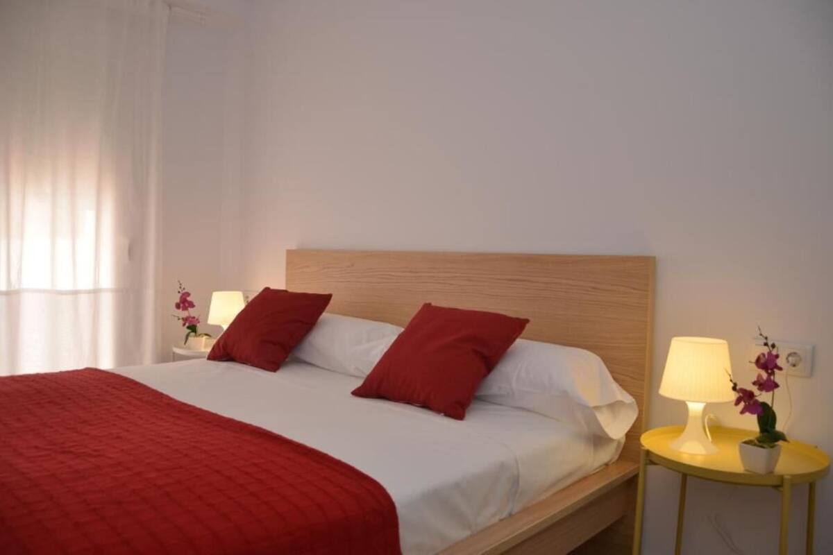 A neatly made bed with a red blanket and pillows, two bedside lamps, and a small table with a flower.
