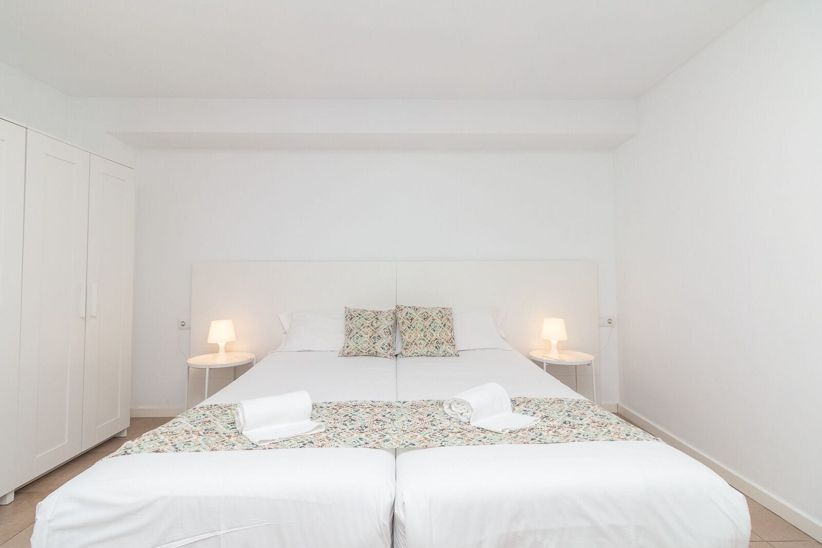 A minimalist bedroom with a double bed, two bedside tables with lamps, and a wardrobe.