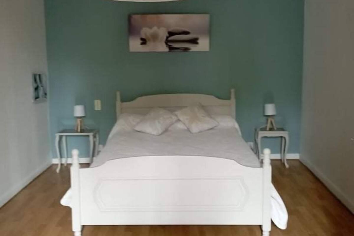 A bedroom with a white bed, two bedside tables, a hanging light fixture, and a wall-mounted artwork.