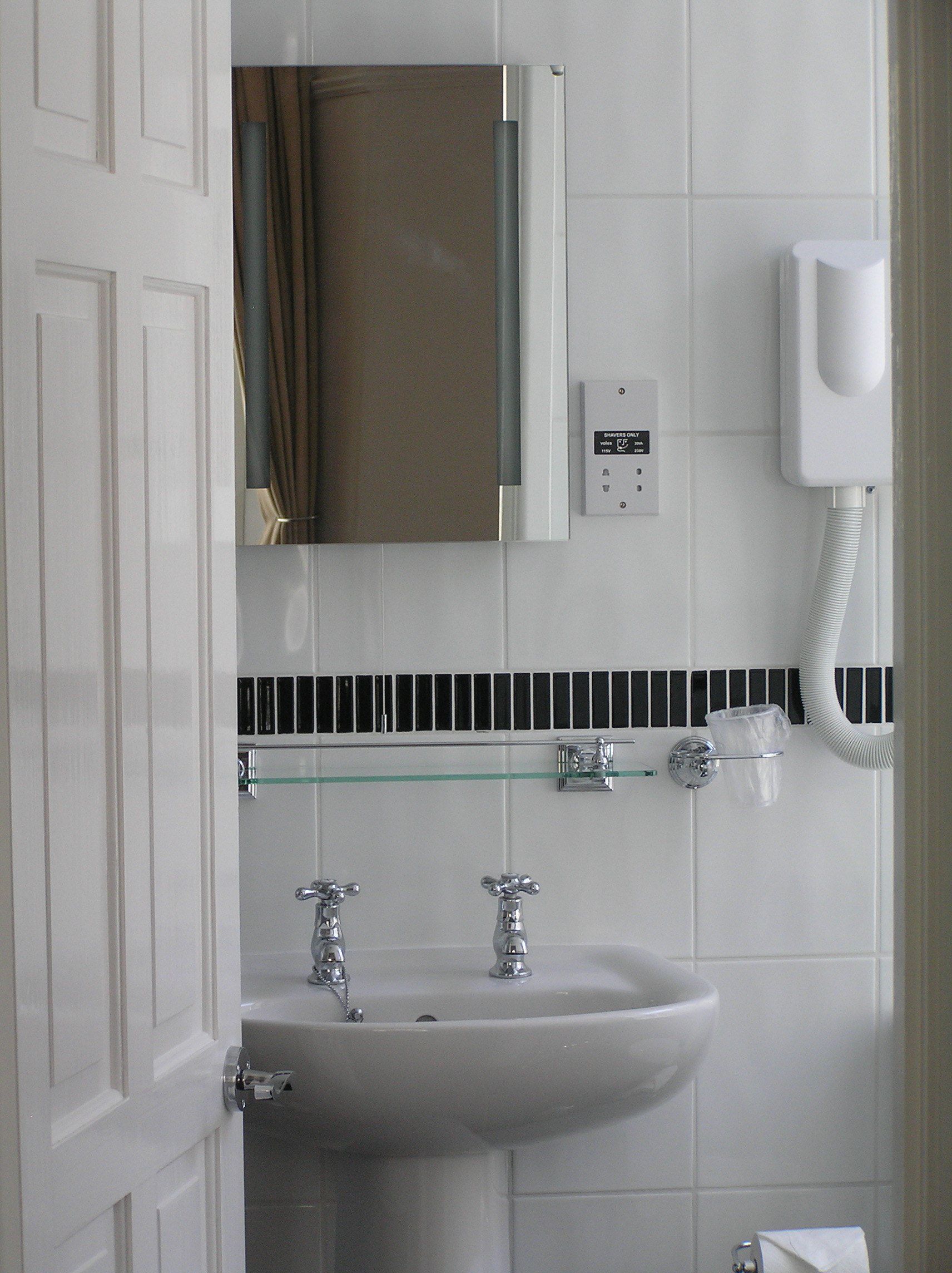 A bathroom with a white sink, two faucets, a glass shelf, a towel rack, a mirror, a towel dispenser, and a telephone.