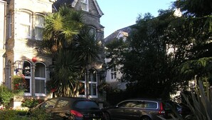 Il s’agit d’un bâtiment historique doté d’une façade en pierre, de grandes fenêtres et d’un toit recouvert de tuiles. Des voitures sont garées devant, et un palmier se trouve sur la gauche.