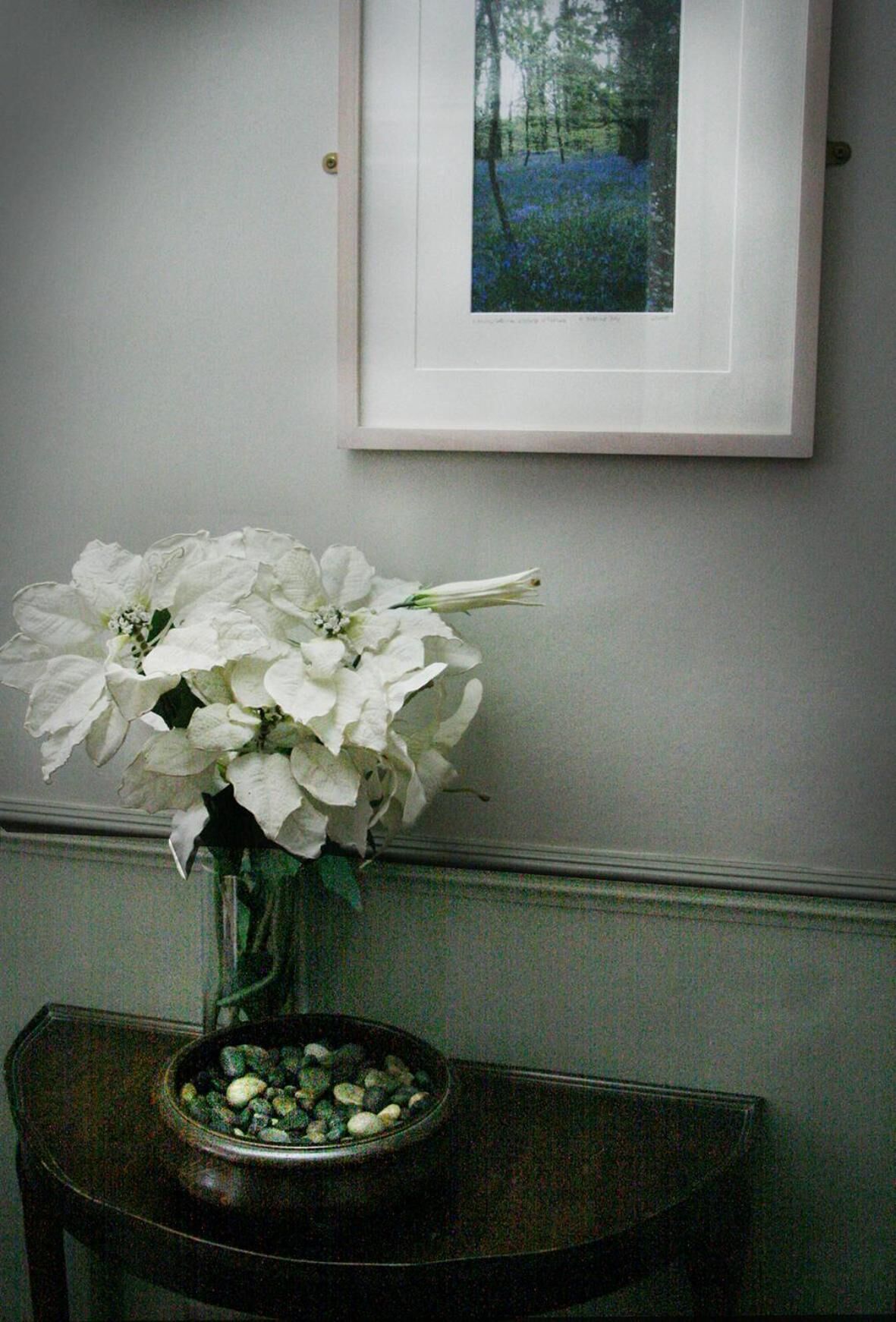 A white vase with white flowers on a dark wooden table, a framed picture on the wall, and a bowl of pebbles on the table.