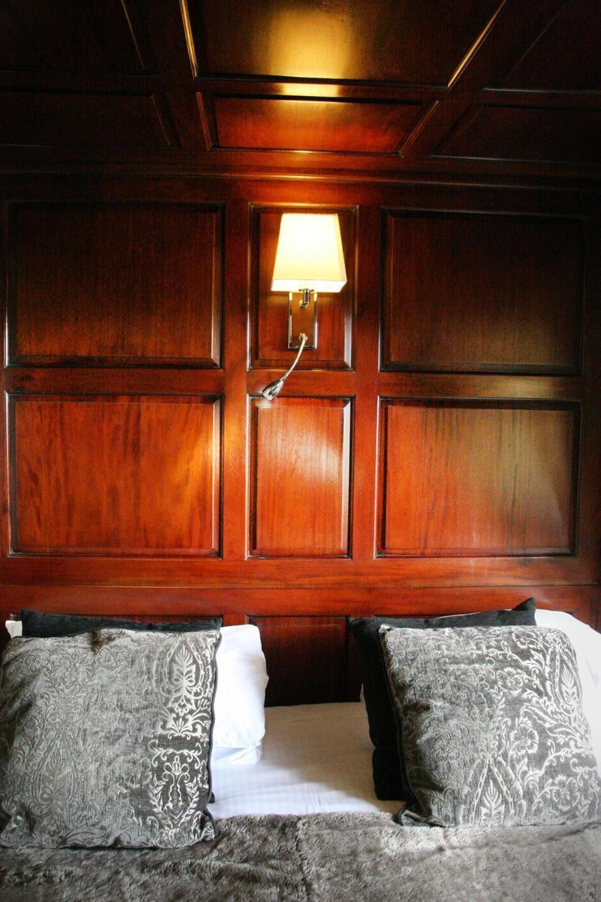 A hotel room with a wooden paneled wall, a bed with patterned pillows, and a wall-mounted lamp.