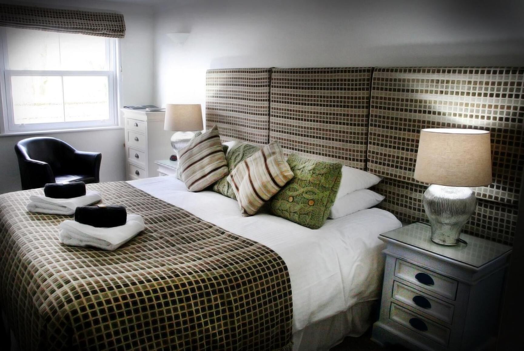 Una habitación de hotel con una colcha a cuadros, un cabecero con diseño de cuadrícula, una lámpara de noche, una silla y una ventana con cortinas.
