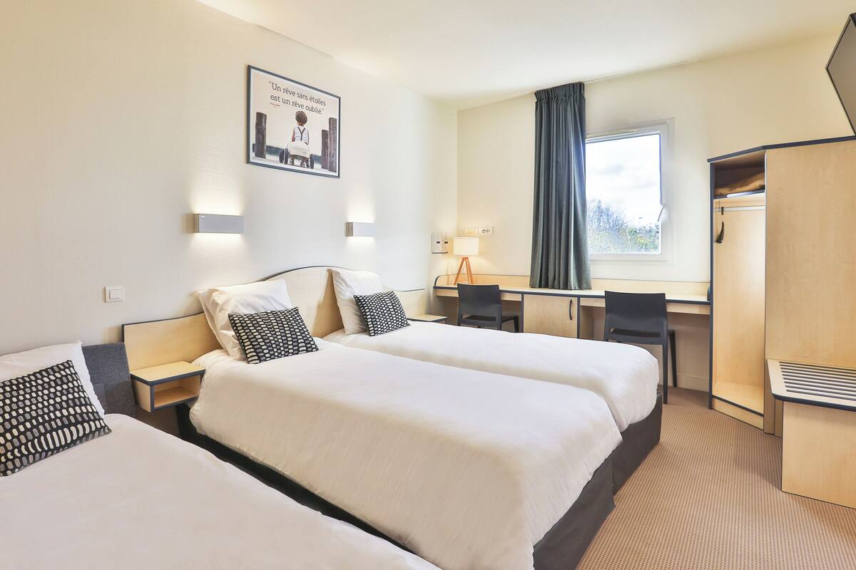 Une chambre d’hôtel avec deux lits, un bureau, une chaise, une armoire et une fenêtre avec des rideaux.