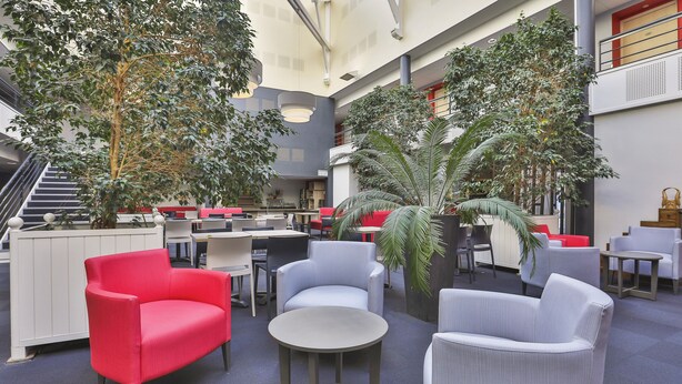 Ruime binnenruimte met zithoek, waaronder een rode stoel, een grijze fauteuil en een ronde tafel. Er staan potplanten en een trap op de achtergrond.