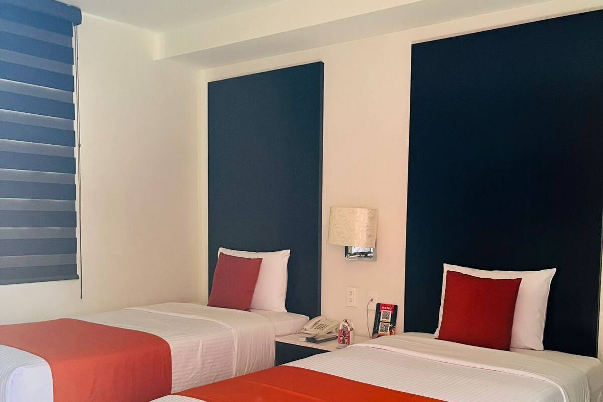 A hotel room with two beds, each with a red and white bedspread, a nightstand with a lamp, and a wall-mounted phone.