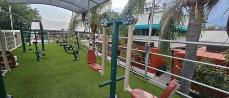 Un gimnasio al aire libre con equipo de ejercicio, una zona cubierta y palmeras.