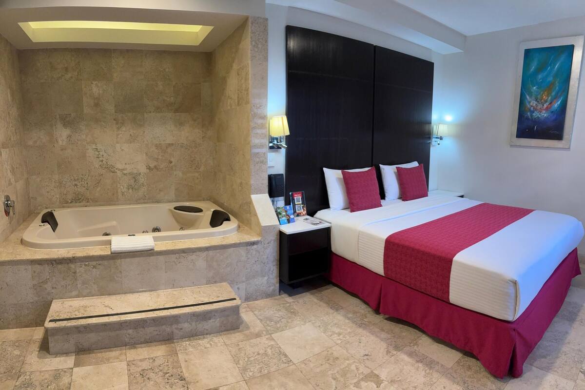 A hotel room with a large bathtub, a bed with red and white bedding, and a painting on the wall.