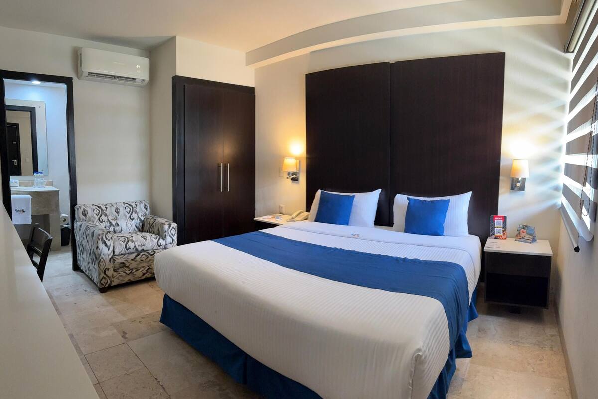 A hotel room with a large bed, a chair, a wardrobe, a desk with a lamp, and an air conditioning unit.