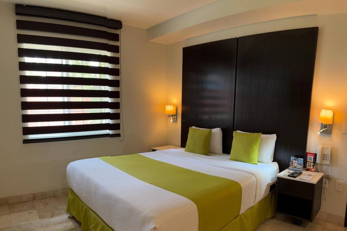 A hotel room with a large bed, a nightstand, a wall-mounted phone, and a window with blinds.