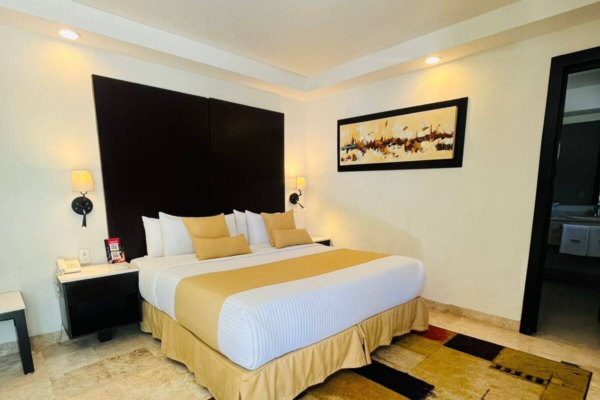A hotel room with a bed, bedside tables, a nightstand, a wall-mounted artwork, and a patterned rug.