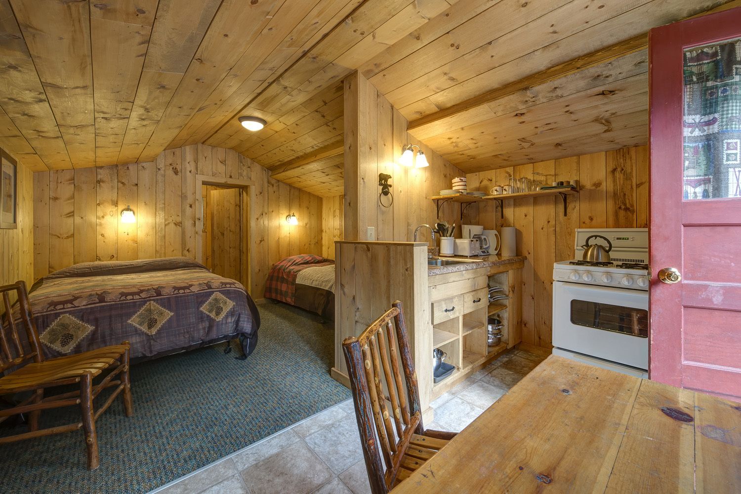 A cozy cabin with a wooden interior, a bed, a desk, and a kitchenette.