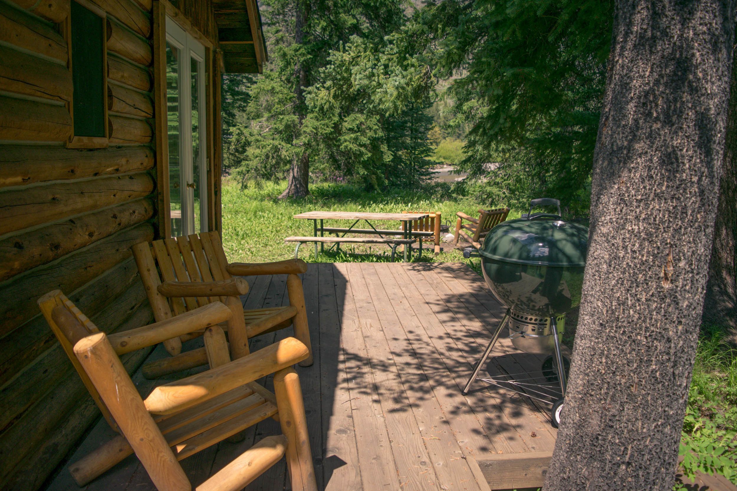 Een houten terras met een picknicktafel en stoelen, een barbecue en een blokhut in een bebost gebied.