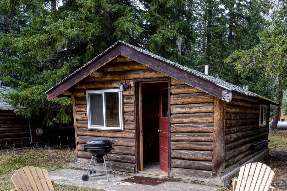 A log cabin with two wooden Adirondack chairs and a metal fire pit in front.