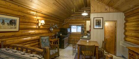 Interior de una cabaña de troncos con comedor, cocina y una zona de estar acogedora con alfombra.