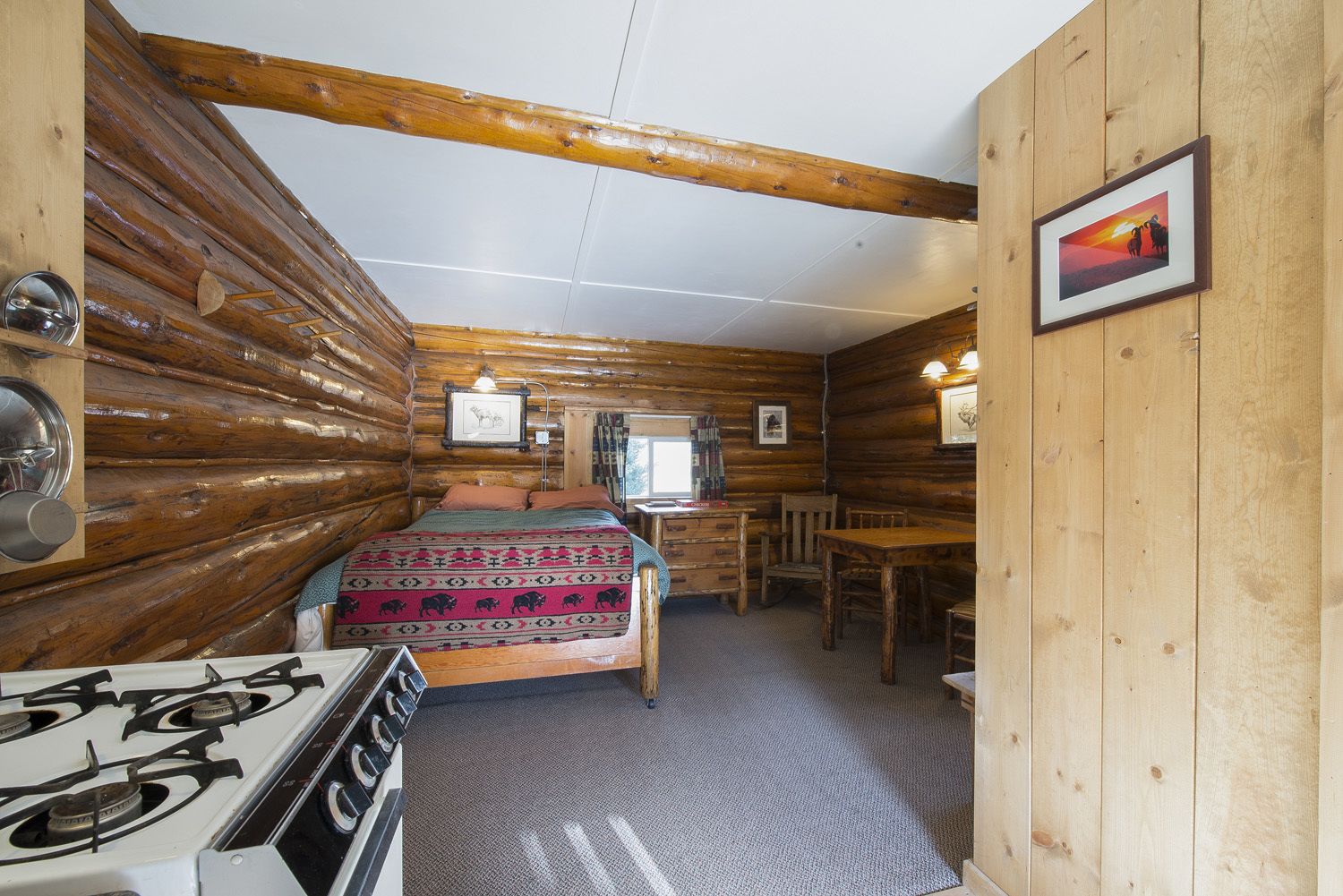 A log cabin room with a bed, a desk, a chair, a stove, and framed pictures on the walls.