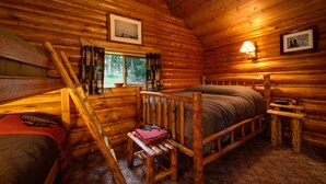 A cozy log cabin room with a bed, a bench, a window with curtains, and a wall-mounted lamp. - Silver Gate Lodging (Silver Gate)