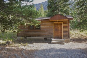 A small log cabin with a wooden door and windows, surrounded by trees and a gravel path.