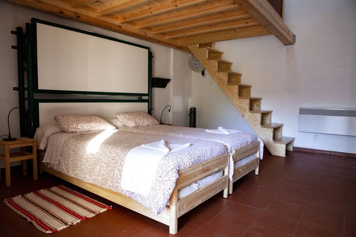 Un dormitorio con una cama de madera, una escalera de madera, una pared blanca y un suelo de baldosas.