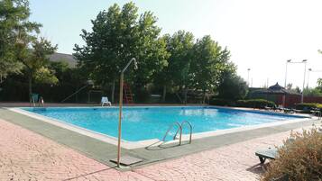 A rectangular swimming pool with a paved deck, surrounded by trees and a covered area with lounge chairs.