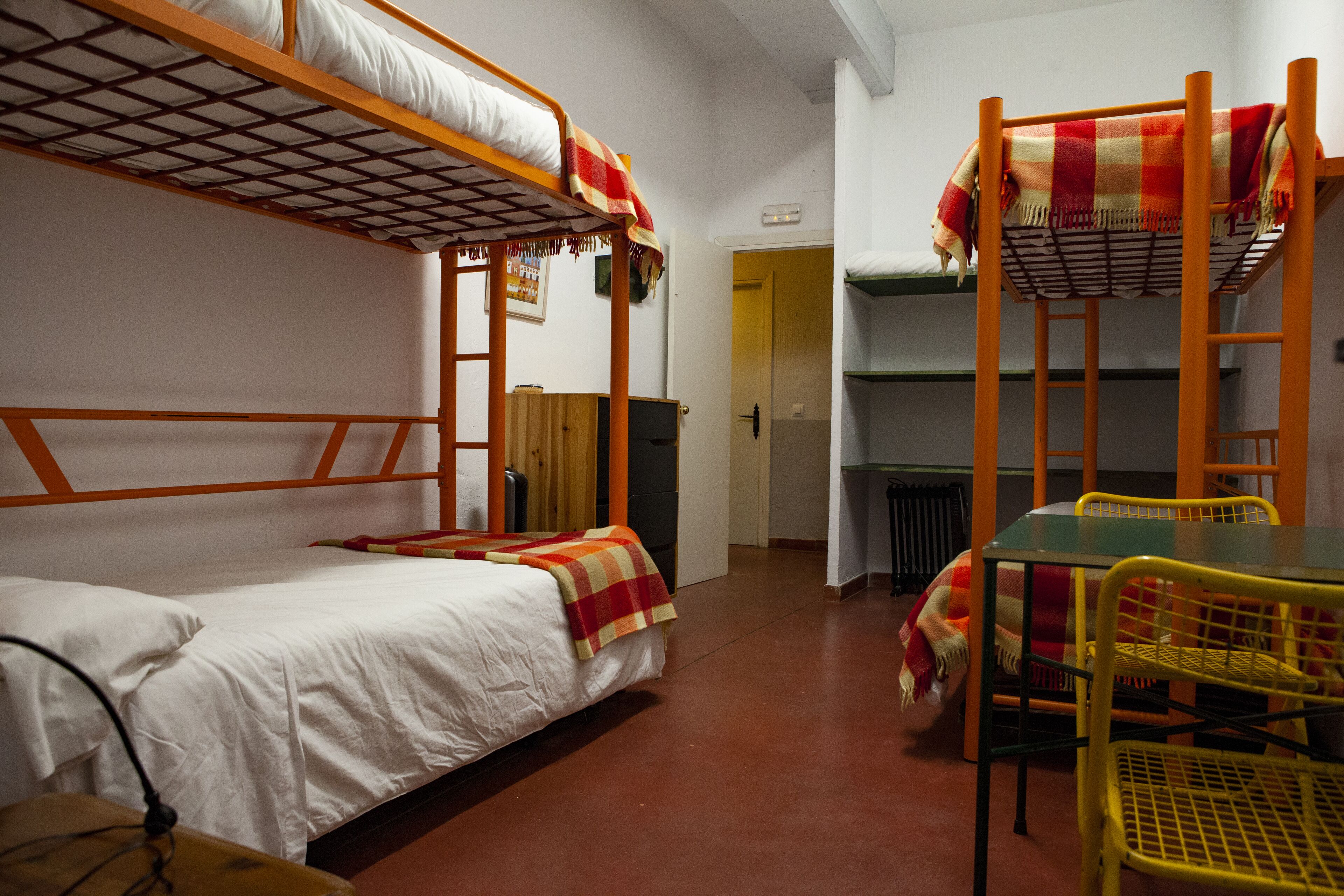 A dormitory room with bunk beds, a table, chairs, and a radiator.