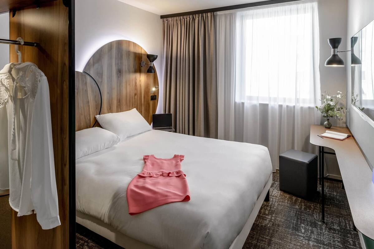 Une chambre d’hôtel avec un lit, un bureau, un ordinateur portable, un miroir et une fenêtre avec des rideaux.