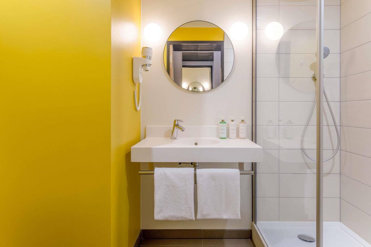 Une salle de bain moderne avec un mur jaune, un lavabo blanc, un miroir, un téléphone et une douche.