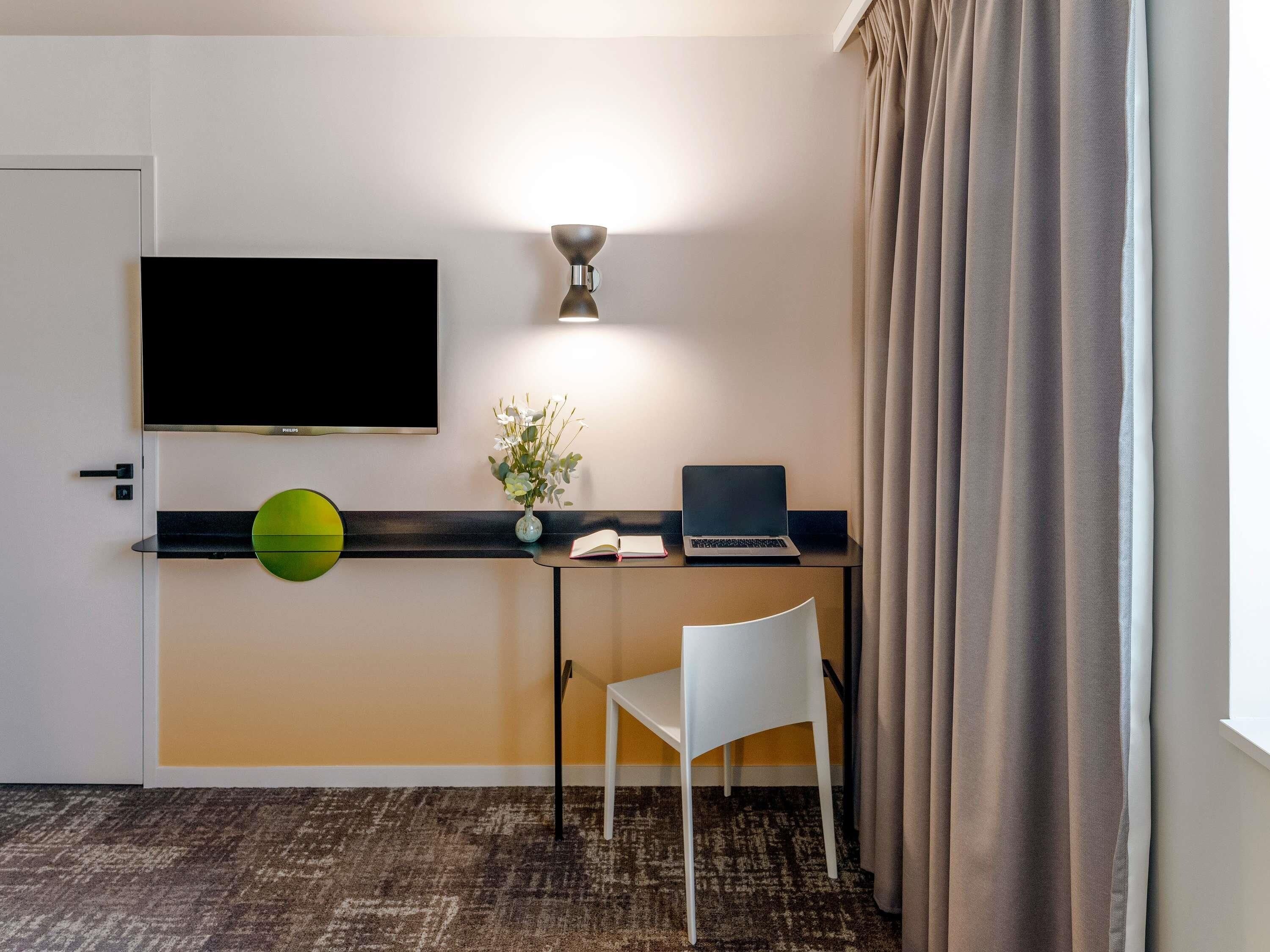 A hotel room with a desk, a laptop, a flat-screen TV, a vase with flowers, a white chair, and a patterned carpet.