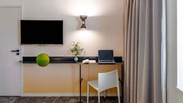 Ein Hotelzimmer mit Schreibtisch, Laptop, Flachbildfernseher, Blumen in einer Vase, einem weißen Stuhl und einem gemusterten Teppich.