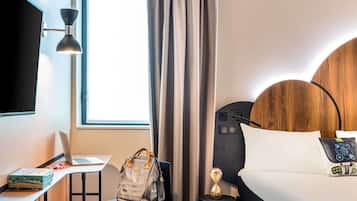 A hotel room with a bed, a desk with a laptop, a chair, a handbag, a clock, and a TV mounted on the wall.