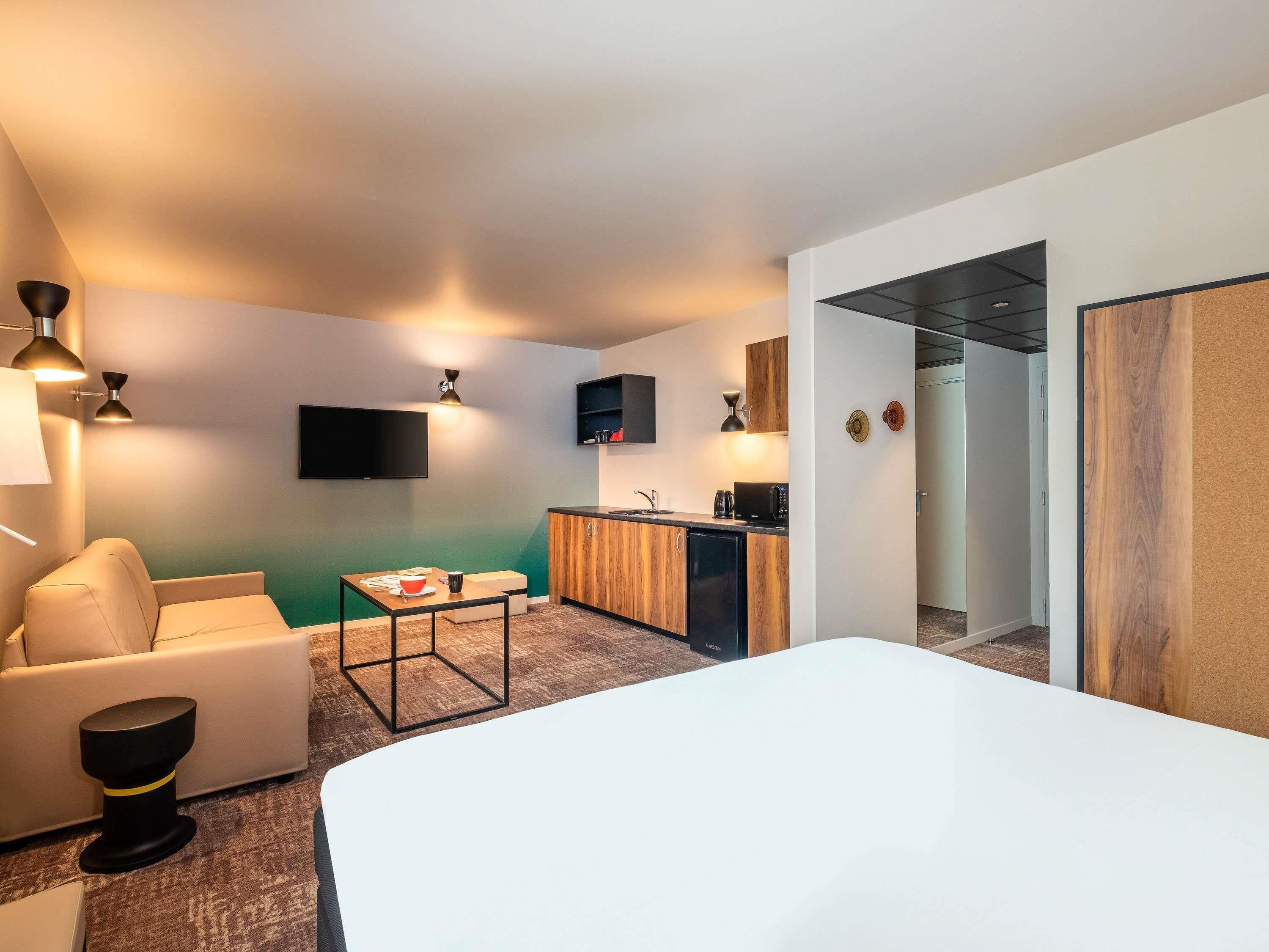 A modern hotel room with a bed, a sofa, a small table, a kitchenette, and a TV.