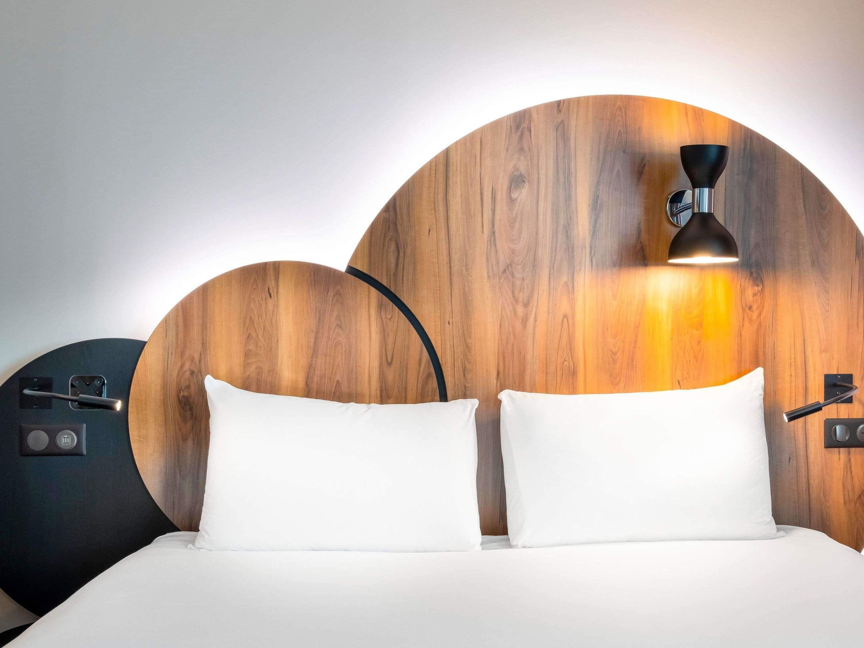 Una cama con ropa blanca, cabecera de madera y una lámpara de pared.
