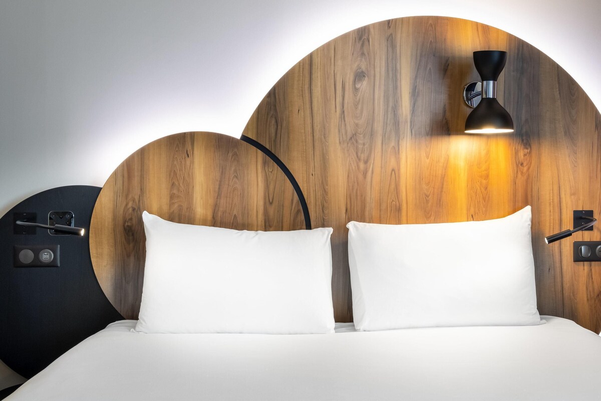 Een bed met witte lakens, een houten hoofdbord en een wandlamp.